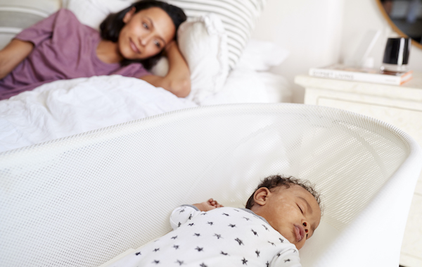 close up of young adult mother lying on her bed looking down at her three month old baby sleeping in his cot