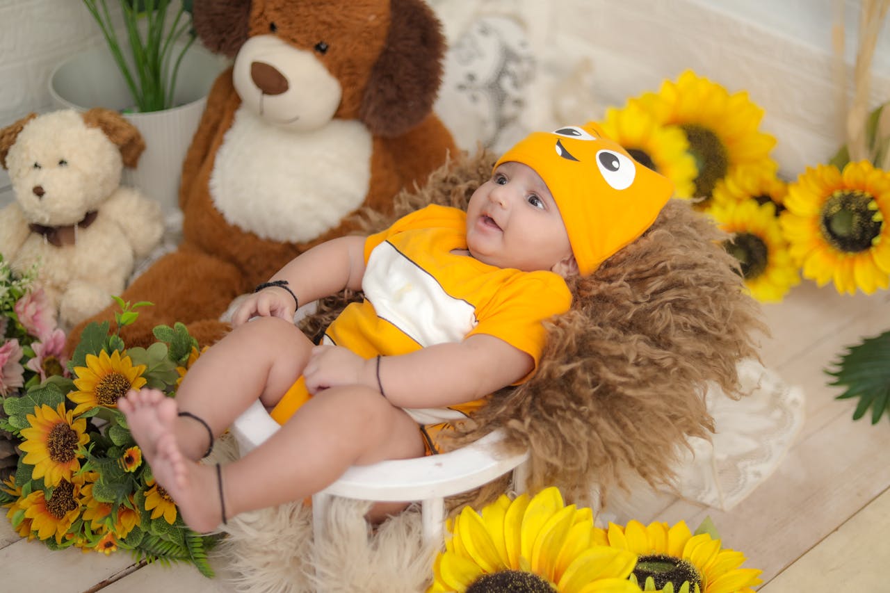 Cute baby dressed in orange outfit surrounded by toys and sunflowers.