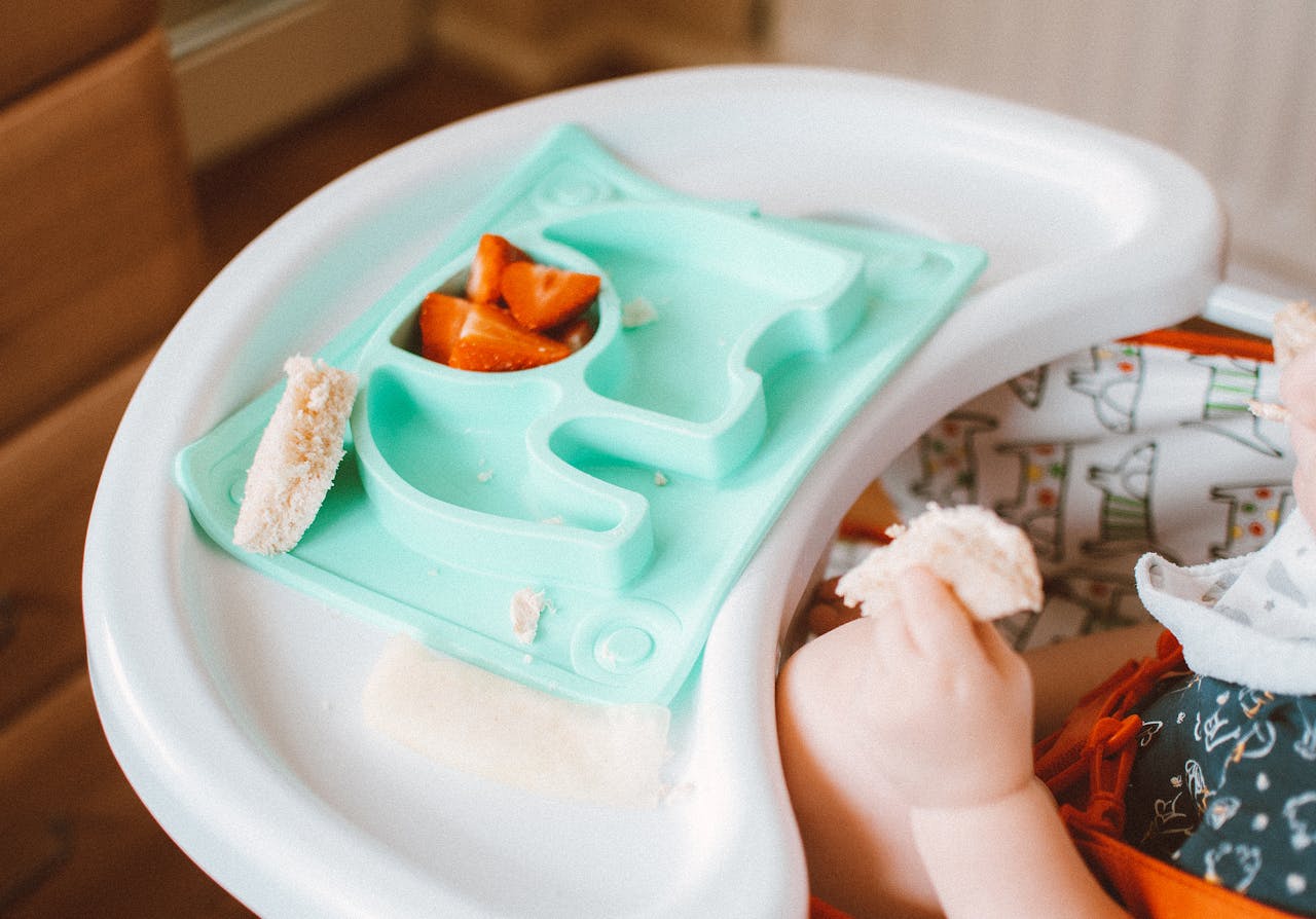 Adorable baby eating bread and carrots in a high chair with colorful tray.
