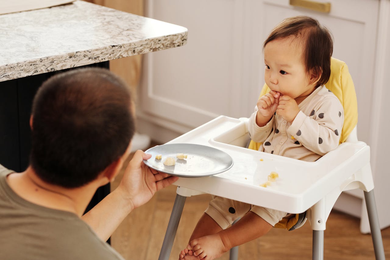 Adorable baby in a high chair eating while a parent offers food, indoors.