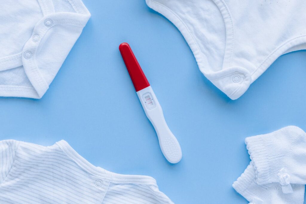 Flat lay of positive pregnancy test surrounded by baby clothes on blue background.