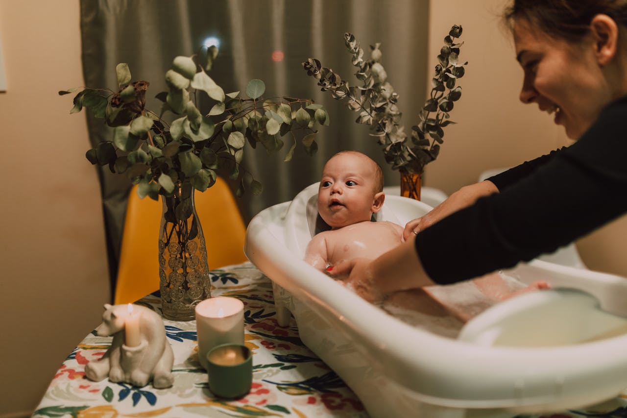 A mother lovingly baths her baby in a small tub surrounded by warm lights and plants.